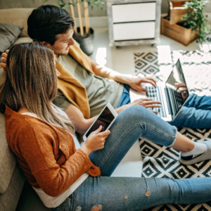 Lady and man sitting on floor. The lady is holding an ipad and the man is holding a portable computer. They appear to be on a video call together.